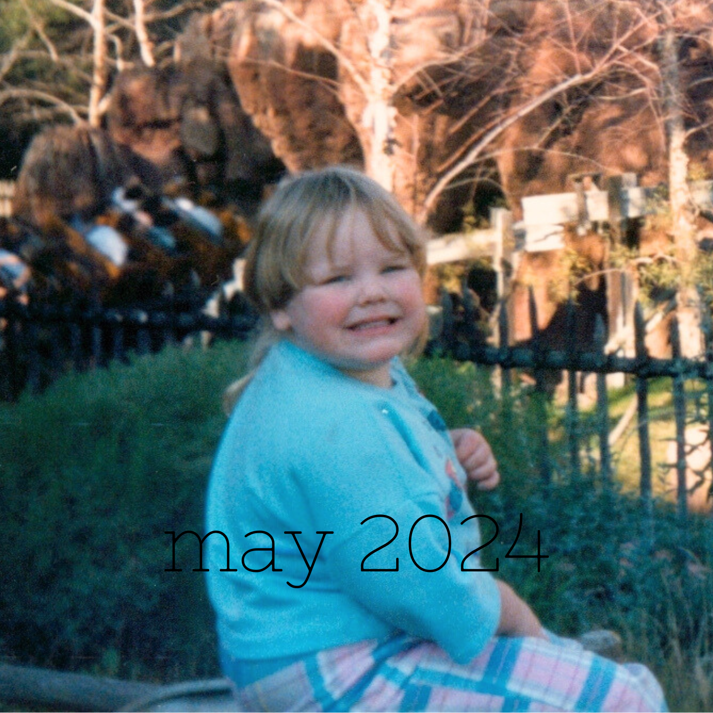 a square image of a four year old kid with the worst mullet of all time sitting in front of big thunder mountain at disneyland in pink and blue plaid pants and a blue shirt while smiling in a grimacing emoji fashion