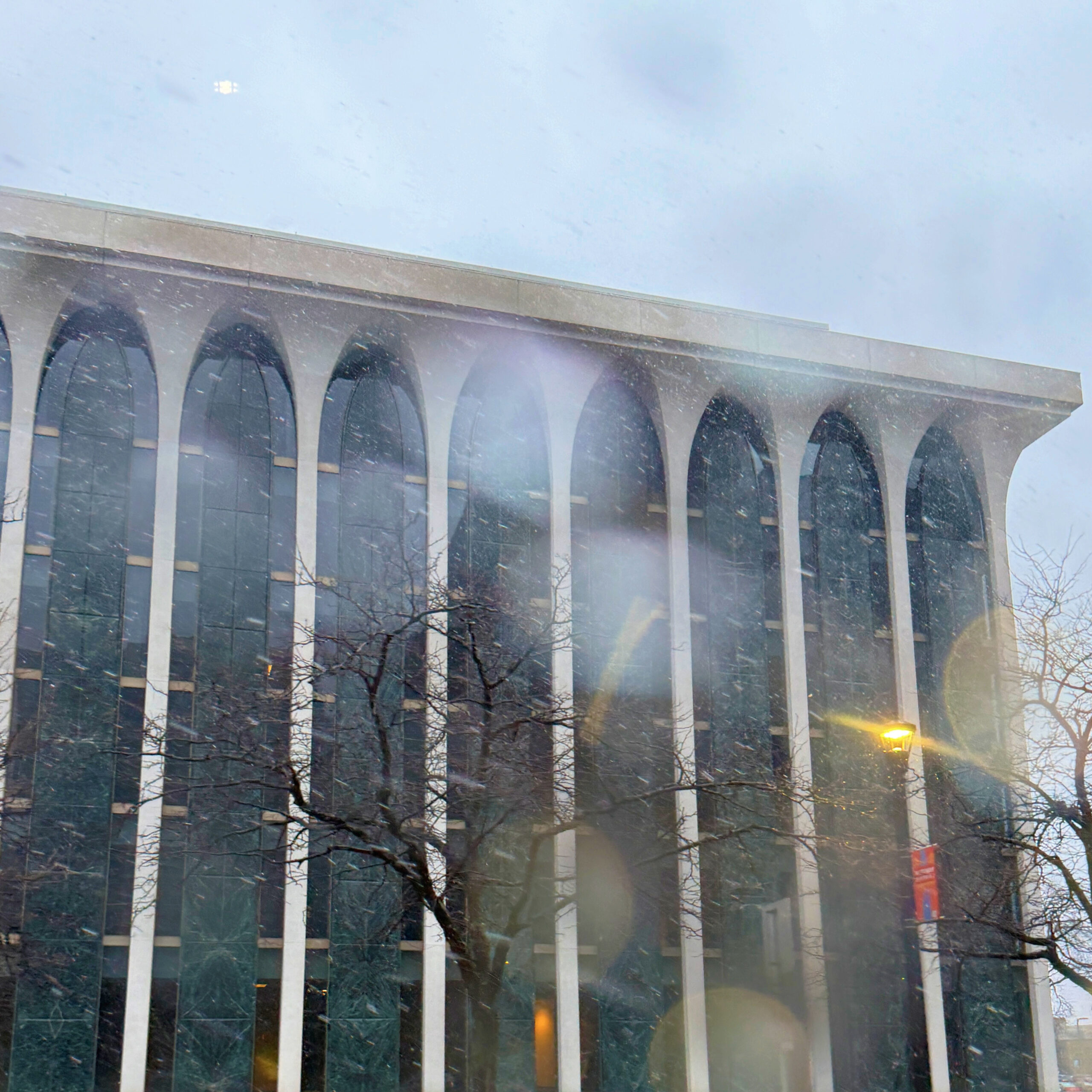 a square image of the corner columns and windows of the northwestern national life building in wet snow, there is light bokeh and streaking from a streelight and bare trees in front