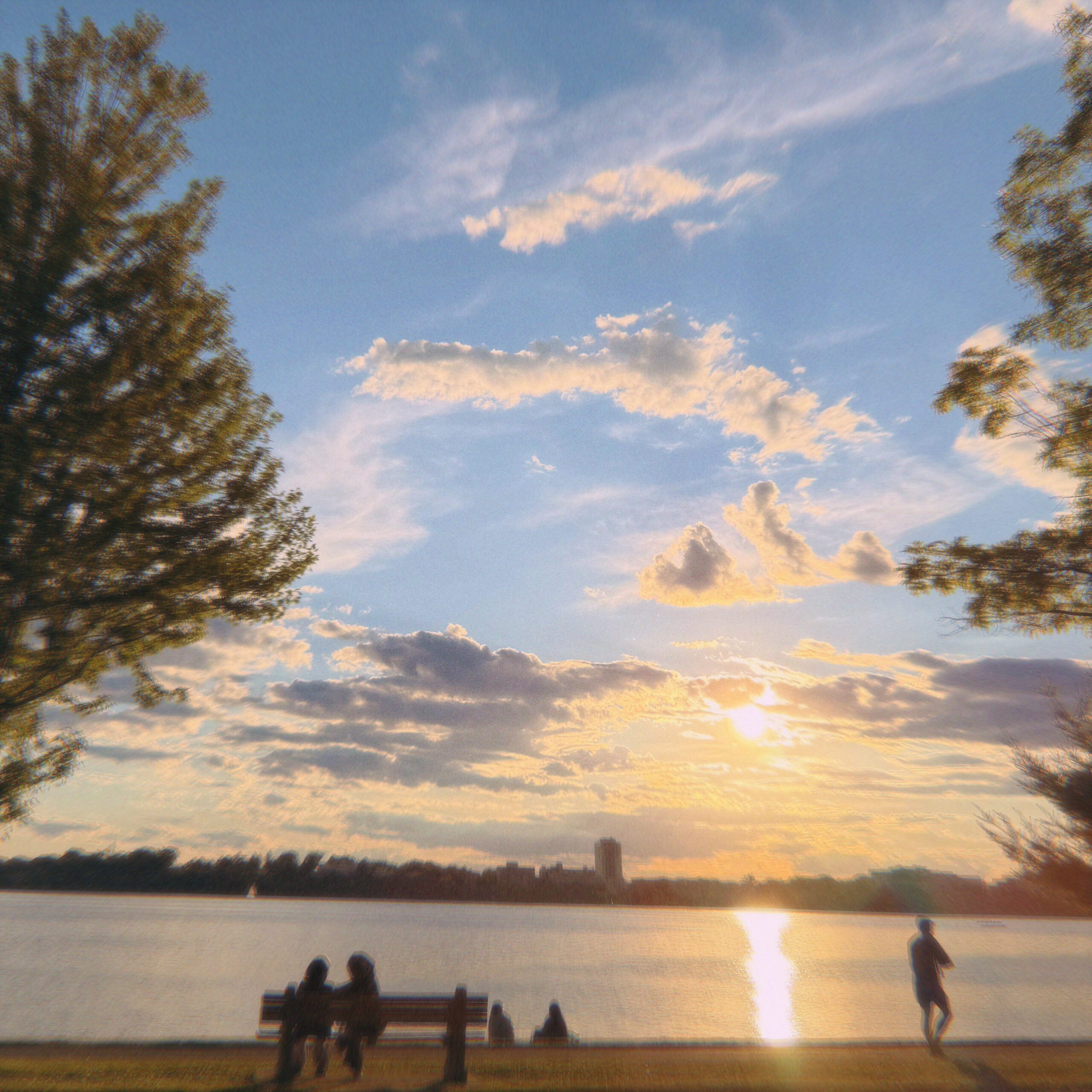 a square image of people alongside a lake, two people sit on a bench and another walks, the sunset makes a strip of light on the water where it shines between fluffy clouds in a blue sky