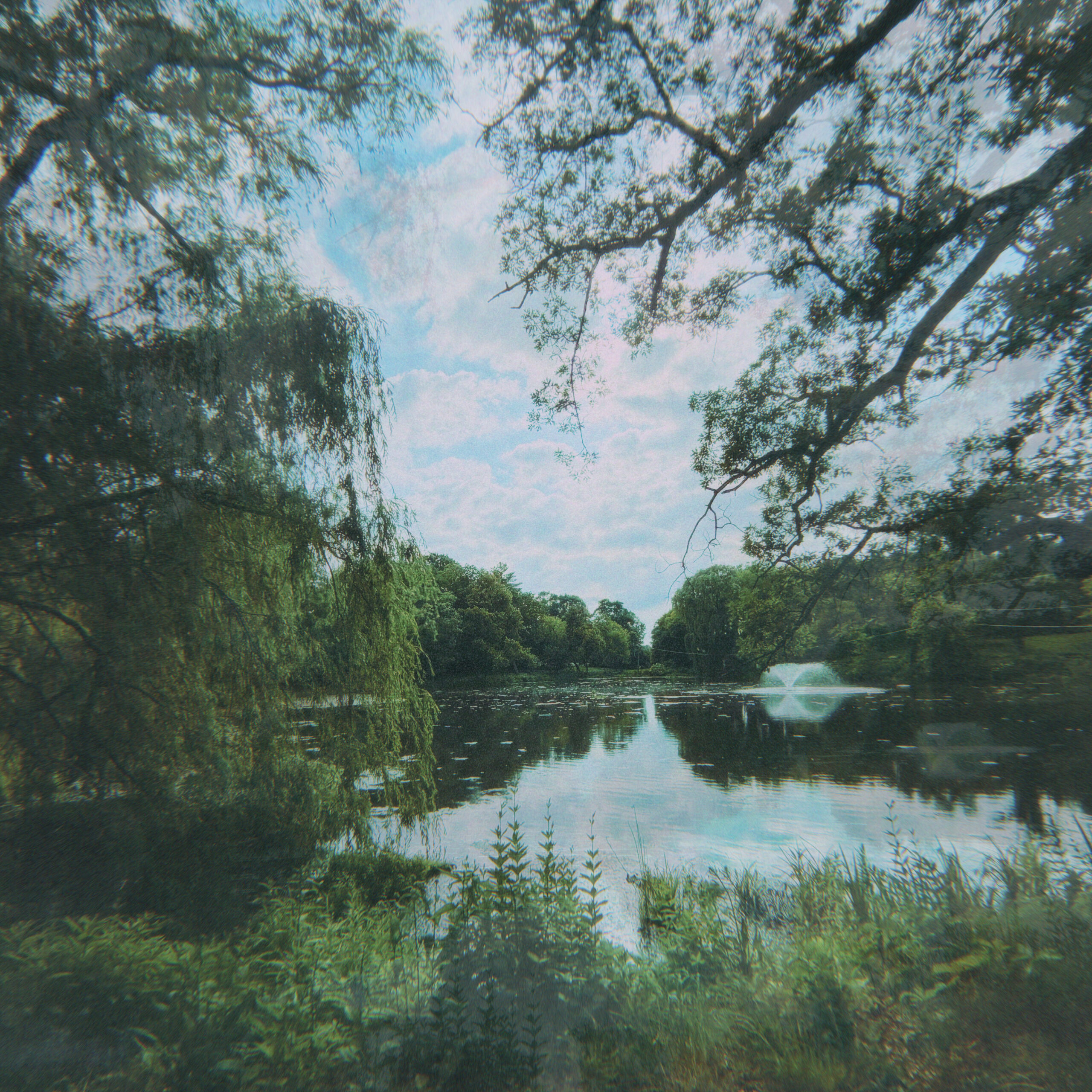 a square image of a section of lake with a spray fountain below blue sky and whiter clouds, the lake is surrounded on all sides by greenery including a weeping willow to the left
